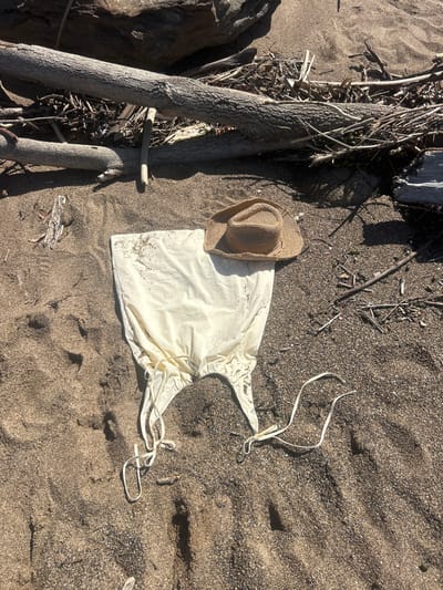 The scene is located on a sandy beach or shoreline in Coalcliff, Australia. A cream-colored, sleeveless garment, possibly a dress or cover-up, with visible drawstrings is laid flat on the dark, gritty sand, displaying numerous fine sand particles across its surface. A light brown woven straw hat rests on top of the garment, positioned slightly right of its center. In the background, several large, weathered driftwood logs and numerous smaller branches are scattered across the sand, interspersed with additional natural debris like twigs and small pebbles. Distinct shadows are cast across the sand and objects, indicating direct overhead sunlight. No individuals are present, and no specific actions are depicted.