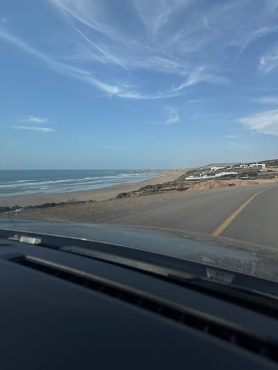 The image shows a coastal road in the city of Aquermoud, Morocco. The road curves to the right, with a yellow line dividing the lanes. To the left of the road is a sandy beach, with gentle waves rolling in from the ocean. In the background, a few white buildings are scattered along the hillside overlooking the coast. The sky is a clear blue with some wispy white clouds. The photo appears to be taken from inside a vehicle, looking out over the dashboard.