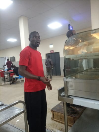 An indoor shot captures a man in a red shirt and black pants, gesturing with his hands. A large, stainless steel food warmer stands beside him, partially obscuring another individual. Background details include tables, chairs, other people, and an exit door. The room is brightly lit, revealing tiled flooring. A serving cart is visible on the left.