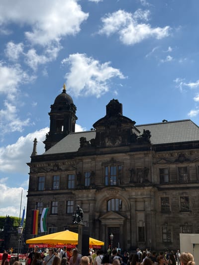 The scene captures the Dresden Frauenkirche in Germany under a partially cloudy sky. The Baroque-style church is visible from a low angle, emphasizing its elaborate architectural details and the golden orb atop one of its towers. In the foreground, a crowd of people gathers, suggesting an event or gathering. Yellow awnings and colorful flags, including what appear to be German state flags, are present. A statue is also visible near the base of the church.