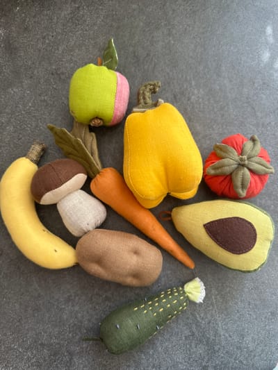 This image displays a collection of handmade plush toys resembling various fruits and vegetables arranged on a dark gray, textured surface. The toys include an apple, a yellow bell pepper, a tomato, a banana, a mushroom, a potato, a carrot, an avocado half, and a cucumber. The background appears to be a flat, possibly stone or concrete surface, with some subtle variations in texture and color. There are no people or actions depicted in the image. The lighting suggests an indoor setting, with soft shadows cast by the objects. The exact location in Chornomorske, Ukraine, is not discernible from the visual information provided.