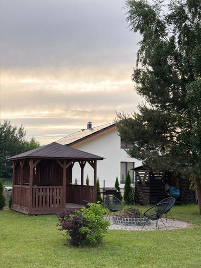 The image depicts a backyard scene in Dreiliņi, Latvia, featuring a white modern house with a dark-tiled roof and solar panels. In the foreground, a dark wooden gazebo with a gabled roof stands on a wooden platform. Adjacent to the gazebo, a circular fire pit area is paved with stones and surrounded by three black wireframe chairs. A pile of firewood is visible in a wooden structure to the right, and a barbeque grill is positioned near the gazebo. Lush green grass covers the ground, with a small bush of dark purple leaves and a green leafy bush in the middle ground. A large tree with green foliage is prominent on the right side of the image. The sky is overcast with soft, diffused light. No people are present in the scene.