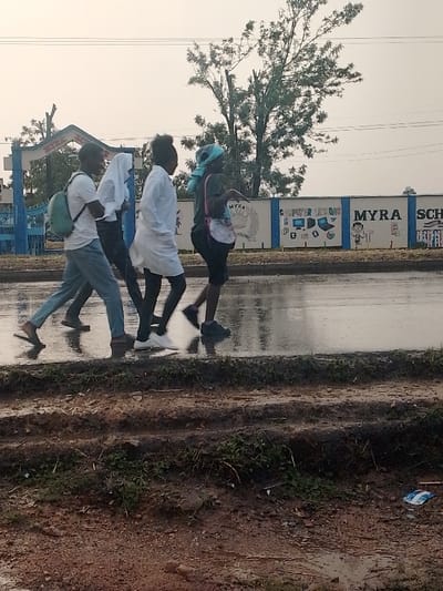 The image shows four people walking across a wet, paved road in front of a school in Isukha ICHINA, Kenya. The school's name, "MYRA SCHOOL," and the phrase "COMPUTER LESSONS" are painted on the blue brick wall. The sky is overcast and the road is wet, suggesting it has recently rained. The people are walking in single file and all have dark skin. Three of the people are women wearing casual clothing. The person in the lead is a man wearing blue jeans and a white t-shirt. He is carrying a green backpack on his left shoulder. He is walking slightly ahead of the second person in line, a woman wearing a white robe-like garment and a white head covering. The third person is a woman in a white blouse and black pants. She is walking with her right arm raised, possibly waving to someone. The fourth person is a woman in a blue scarf and dark clothing, wearing a white backpack. She is walking with her right arm raised, possibly waving to someone. The environment is urban and there is a large, leafy tree in the background.