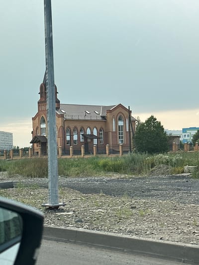 This image captures a segment of a street scene in Abakan, Russia, oriented vertically. The primary focus is a multi-story building with a distinctive arched window design, constructed with reddish-brown brick. The building appears to be residential or a complex of apartments, with several balconies and visible windows. Adjacent to the building, there is a stretch of gravel and sparse greenery, suggesting a roadside environment. A modern-looking street barrier or railing runs across the foreground, partially obscuring the lower section of the building. In the background, a large green tree stands prominently, and further beyond, another building with a lighter facade and blue accents can be seen. The sky is overcast with a pale, light blue hue, indicating a cloudy or hazy day. The overall lighting suggests it is either early morning or late afternoon, given the soft light. No people are visible, and the scene is quiet and devoid of specific activity. The composition implies the photograph was taken from a moving vehicle, possibly looking out from a passenger window, given the angle and the presence of a car mirror in the lower right corner. The location cues, such as the architecture and the overall impression of the setting, align with a suburban or newly developed area in Russia.