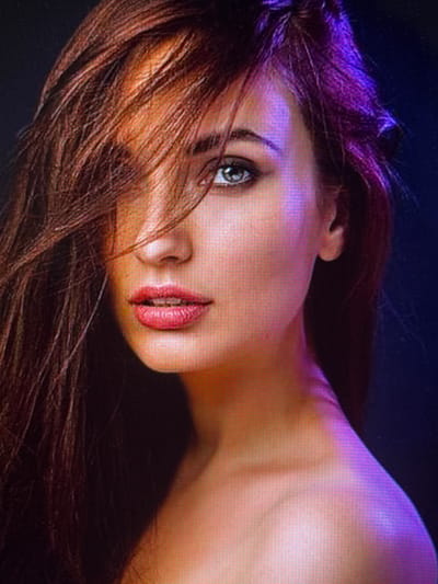 The image is a close-up, side profile portrait of a young woman with long, dark brown hair that is partially obscuring her face. Her eyes are a striking blue-grey, and she is wearing pink lipstick. The lighting is dramatic, with a warm orange glow on the left side of her face and neck, transitioning to a vibrant purple light on the right side of her head and shoulder, against a dark background. There is no discernible location in Taipei, Taiwan, or any contextual environmental details that would indicate a specific city. The focus is solely on the woman's facial features and the artistic lighting.