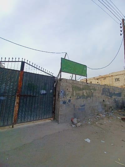 This image depicts an outdoor scene in the city of Kafr Sousa Municipality, Syria, focused on a gateway and an adjacent wall under an overcast sky.
In the foreground, the ground transitions from a lighter concrete/asphalt surface on the left to a dirt and debris-strewn area on the right, littered with various pieces of trash including paper and small rocks.
On the left, a large, dark-colored, industrial-style metal gate, approximately two meters tall, is partially open. The gate features a grid pattern and an ornate, spiked top section. Rust is visible on the gate's frame and hinges. The gate is connected to a sturdy concrete pillar on its right side.
To the right of the gate, extending further right across the midground, is a low, unpainted concrete block wall, roughly constructed and showing significant weathering, discoloration, and grime. Patches of darker stains and possibly moss are present on its surface.
Mounted above the concrete wall and pillar, a rectangular green sign with white or light-colored Arabic text (mostly illegible due to resolution and angle) is visible. The sign is supported by metal poles and is angled upwards slightly.
In the background on the right, a two-story building constructed of light-colored masonry (possibly stone or concrete blocks) is visible. It features multiple windows and appears to be in a state of disrepair or aged. Further left in the background, behind the open gate, another structure made of similar materials is partially visible.
Overhead, a network of utility wires spans the scene. A dark brown wooden utility pole with several crossarms and multiple power lines runs vertically along the far right edge of the frame. A prominent dark wire sags horizontally across the upper-middle section of the image. The sky is a uniform, bright grey, indicating an overcast day. No individuals are present in the scene.