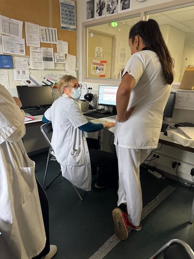An administrative or workstation area within a professional or medical facility in Tourcoing, France, is depicted. Three individuals are present. A woman with light hair, wearing glasses and a light blue surgical mask, a white lab coat over a blue long-sleeved shirt, and dark pants, is seated on a black folding chair at a desk. She faces towards the right of the frame, with her hands on a document in front of a black computer monitor displaying data with red lines. To her left, another black computer monitor is visible but dark. A second woman, with dark hair in a high ponytail, wearing a white scrub top and pants, stands with her back largely to the camera, facing the seated woman. Her right hand rests on her hip, and she wears red and brown flat shoes. A third individual, partially visible on the far left, wears a white lab coat or scrubs over dark clothing and white shoes. The desk is cluttered with papers. A cork board behind the desk is covered with various documents, charts, and diagrams, some with French text including "OPTIMANEW." Above the cork board, framed black and white photographs are displayed. A glass partition behind the seated and standing women features a yellow sign with black pictograms and French text, including "RÉSERVÉ PERSONNEL" (Reserved Personnel) and "CHAPITRE ACCIDENT DU TRAVAIL" (Work Accident Chapter). The floor is dark green/grey, traversed by a strip of silver tape. Electrical outlets and cables are visible on the wall beneath the desk on the right.