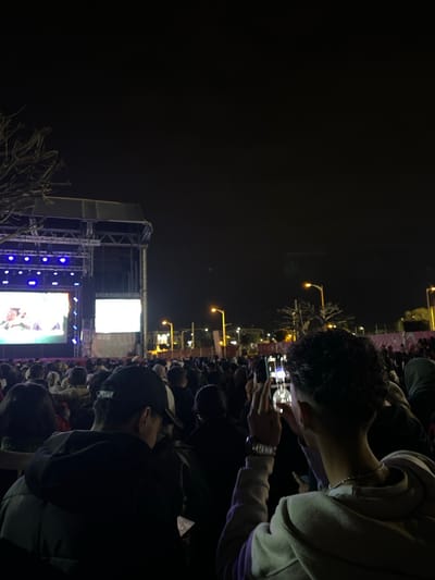 A nighttime view captures a crowd gathered before a stage, illuminated with blue lights and large screens displaying content. Attendees hold up their phones, seemingly recording or photographing the event. Overhead, street lamps cast a soft glow. The overall atmosphere suggests a concert or festival setting.