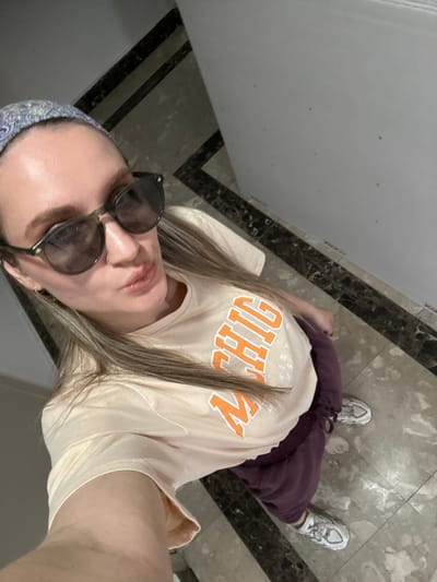 A woman is seen in a selfie taken from a high angle, likely indoors on a marble-tiled floor. She is wearing sunglasses, a light-colored t-shirt with orange lettering, and maroon sweatpants, and has on a patterned headscarf. White sneakers are visible on her feet. The background suggests the location is inside a building, likely in the city of Esenyurt, Turkey, due to the style of tiling seen on the floor.