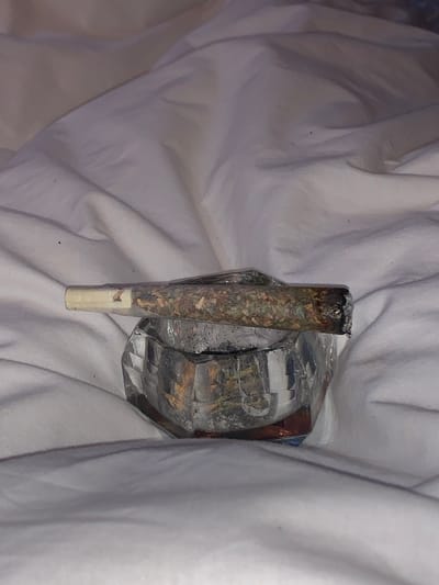 A lit joint rests on a clear, multifaceted ashtray atop a white, wrinkled sheet. The joint, displaying a burnt tip, reveals visible plant matter inside the rolling paper. The ashtray, seemingly used, contains ashes and debris. The wrinkled bedsheet provides a soft backdrop to the scene.
