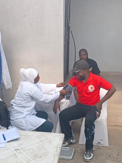 The image depicts a health screening or medical check-up taking place in a semi-outdoor setting, possibly against a building in Tarauni, Nigeria, during the daytime. The central activity shows a woman, dressed in a white lab coat and a white hijab, crouching to operate a blood pressure monitor on the left arm of a seated man.

The man, who appears to be the patient, is wearing a red polo shirt, a black baseball cap, and dark pants with abstract designs. He is seated on a white plastic chair and has a relaxed, smiling expression. His red polo shirt prominently displays a circular logo with the text "EKRA YOUTH FELLOWSHIP" and "LYC WEST" below it. On the ground next to his feet, a grey weighing scale is visible. His sneakers have white crosses and other designs.

Behind the man getting his blood pressure checked, another man in a black t-shirt is observing the process. To the left of the scene, a white plastic table holds an open notebook with handwritten notes and a blue pen. A black backpack is placed near the table. A blue and white piece of cloth hangs on the far left. The background is a plain, light-colored wall. The lighting suggests fair weather conditions during the day.