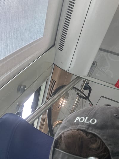 The image depicts the interior of a train car. A person wearing a grey baseball cap with "POLO" embroidered on it is visible from behind. The cap partially obscures a person's hair. In the background, a bicycle is mounted on a rack. The train car features a window with a textured blind, white interior paneling, and a metal railing. The scene is lit by natural light coming through the window and interior lights. There is no visible indication of the specific location within the city of Salses-le-Château, France.