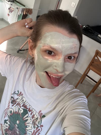 The image is a selfie taken indoors, likely in a home. The primary subject is a young woman wearing a face mask. She has dark hair, brown eyes, and is sticking her tongue out playfully. Her face is partially covered in a light green clay mask. She is wearing a light-colored t-shirt with a graphic print of butterflies and text, which is partially visible. In the background, a wooden chair and a tiled floor are visible, suggesting an indoor setting. The lighting suggests it is daytime. There are no clear indications of the weather or specific time of day beyond daytime. The overall mood of the image is casual and lighthearted, conveyed by the woman's playful expression. There are no other people clearly visible in the frame, though a blurry image of feet could indicate someone else is present. The location context of Limassol, Cyprus, does not provide any immediately discernible visual cues within the image itself.