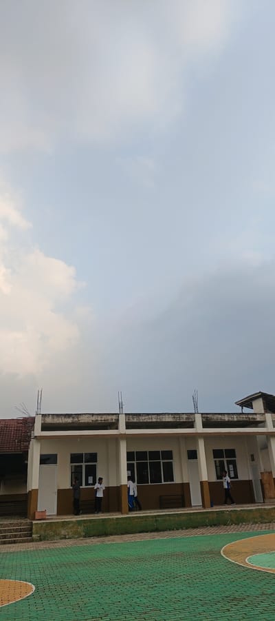 The image depicts an outdoor scene in front of a white and brown school building, likely located in Sukabumi, Indonesia, given the context. The building has a covered walkway supported by white columns, with large windows. Two visible metal rebar protrusions extend from the top of the building's facade. In front of the building, a green paved area, possibly a court, is visible. Several individuals, appearing to be students in casual attire, are present: one is standing near a white door on the left, two are near windows in the center, and two more are walking along the covered walkway on the right. One student is wearing a white shirt and blue pants, while others are in lighter colored shirts. The sky above is overcast with a mix of lighter and darker clouds, suggesting potential for rain or a gloomy day. The overall impression is a snapshot of a school environment during what appears to be an ordinary day.