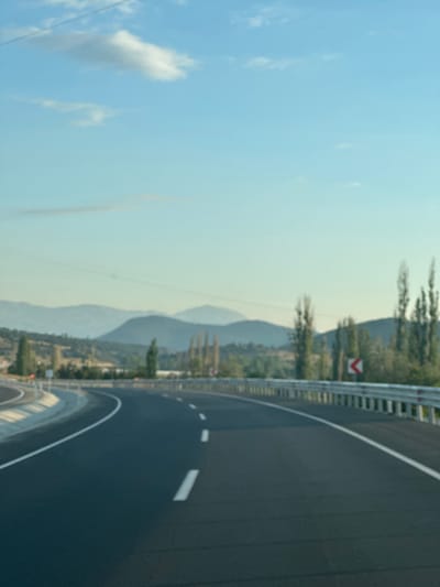 The image depicts a road curving through a mountainous landscape under a blue sky with scattered clouds. The road is paved with asphalt and marked with white lines. A metal guardrail runs along the right side of the road, with a red and white directional sign indicating a curve to the right. On the left side of the road, a concrete barrier is visible. In the background, rolling hills and mountains are layered, with sparse trees and vegetation dotting the landscape. The overall scene suggests a rural or semi-rural setting, possibly a highway or a scenic route. There are no people or specific urban elements visible. The image appears to be taken from a moving vehicle, indicated by the perspective and slight blur. The location is identified as Seydikemer, Turkey.