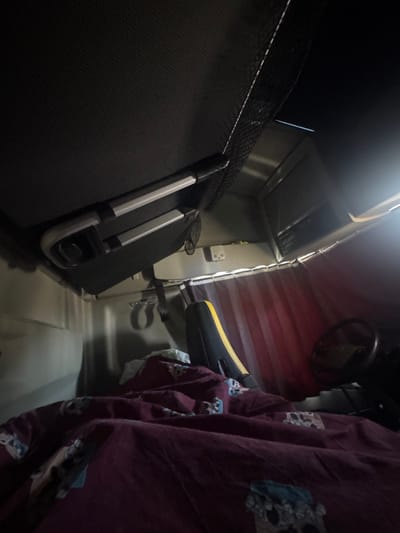 The scene depicts the interior of a vehicle cabin, located in Tortoreto, Italy, under low-light conditions, likely at night. The foreground is dominated by a rumpled maroon duvet with a repeating pattern, covering what appears to be a sleeping bunk or bed. A white or light-colored pillow is partially visible beneath the duvet. To the right of the duvet, a vehicle seat's backrest, featuring black upholstery with a prominent yellow stripe, is discernible. Further to the right, a steering wheel is partially visible, indicating the vehicle's cockpit area.

Above the bed, a collapsible, gray and silver rectangular object, possibly a table or shelf, is secured to the ceiling. Above this object, a black mesh storage net is stretched across the ceiling. Along the far wall, below an overhead compartment or appliance (possibly a small television or microwave oven), red-toned curtains with vertical stripes are drawn across a window or opening. A white power outlet with multiple sockets is mounted on the light-colored wall to the left of the curtains. The overall environment is enclosed and functional, consistent with a truck sleeper cabin or a camper van interior. No individuals are present or visible in the image.