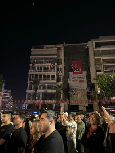 The image shows a gathering of people in the evening in İzmir, Türkiye, at the coordinates provided. The crowd is standing in front of a building that is partially obscured by a large banner. The banner is red and white with the letters CHP in large, bold letters, followed by the text "Aday yarımada sandığı bizimde İSTİYORUZ." The banner is hanging from the building's second floor, and there are several Turkish flags draped across the building. The crowd is facing the building, and many are holding their cell phones up, possibly taking photographs or videos. There are palm trees visible in the background, and the street is lined with buildings, some of which are lit up. The scene appears to be a political rally or demonstration. The crowd is diverse in age and gender, with a mix of men and women. The people in the foreground are close to the camera, with some faces obscured by others. The scene is lit by streetlights and the ambient glow of the city lights, which creates a soft and hazy effect.
