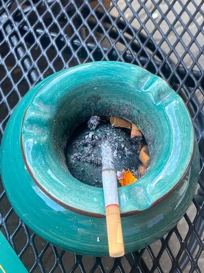 The image shows an outdoor scene, likely on a patio table, with a green ceramic ashtray filled with cigarette butts and ash. A single cigarette rests in the ashtray, with its filter visible and the burning end partially obscured by ash. The ashtray is situated on a black metal mesh surface. The background is out of focus, but appears to be a continuation of the mesh surface, suggesting an outdoor furniture setting. The lighting suggests it is daytime, with natural light illuminating the scene. There is no indication of specific location within Lütjenwestedt, Germany, or any people present.