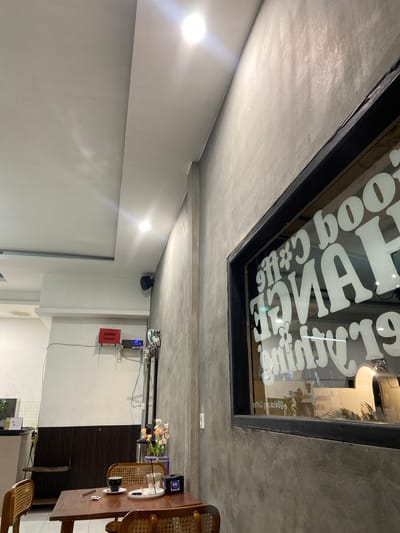 This image depicts the interior of a cafe, likely in Sekar Mawar, Indonesia. The scene is dominated by concrete and white walls, creating a minimalist and modern aesthetic. A large window with white text on it is a prominent feature, reflecting light and showing a glimpse of the outside. The text appears to be a logo or name for the establishment, with stylized lettering and floral elements.
In the foreground, a wooden table with chairs is set up, suggesting a dining or seating area. On the table, there is a coffee cup and saucer, a small vase with flowers, and what looks like a device with a blue light, possibly an electronic gadget. The lighting is a mix of bright artificial lights and natural light filtering in, creating reflections on the surfaces. The overall ambiance suggests a quiet, perhaps evening or late afternoon setting, as indicated by the indoor lighting. There are also some visible electronic devices mounted on the wall, including a red box and what appears to be a security camera. The image is taken from a slightly angled perspective, adding depth to the scene.