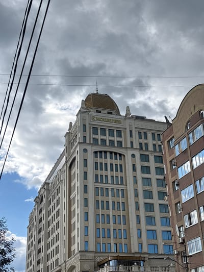 The image shows the "Koshelev" residential complex in Novokuybyshevsk, Russia, on an overcast day. The complex is a tall, multi-story building with a beige facade and a distinctive golden dome. The building features numerous windows, some with blue tinted glass, and decorative architectural elements. To the right, another building with a brown facade and balconies is partially visible. Overhead, black electrical wires crisscross the sky, contrasting with the grey clouds and patches of blue sky. The perspective is from below, looking up at the buildings. No people are visible in the image.