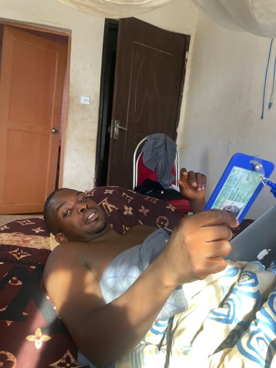 A man is lying on a bed in a room. He is holding a blue identification card in his right hand. A laptop is next to him on the bed. The bed has a brown and gold Louis Vuitton patterned comforter and a white and blue patterned duvet. A gray shirt is on a red and chrome chair. A dark brown door is in the background. The scene likely takes place in a residential interior. Specific location within Rafin Bauna, Nigeria, cannot be determined from this image.
