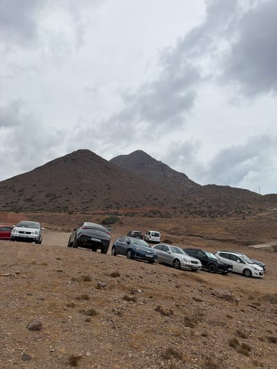 This media file depicts a parking lot situated on a rocky, arid hillside with a mountain in the background. The scene is set under an overcast sky, suggesting a cloudy day. A line of several cars is parked diagonally across the frame, suggesting they are parked on a slope. The cars are of various models and colors, including white, black, and silver. Some of the cars have visible license plates.

The overall setting is natural and undeveloped, with sparse vegetation. The mountain is covered in dry, rocky terrain, characteristic of a desert or semi-arid environment. The sky is filled with grey clouds, indicating potentially cool or damp weather. There is no indication of specific activity or event beyond cars parked in what appears to be a designated or informal parking area. The lighting is diffused due to the clouds, with no strong shadows.

Visible text on license plates includes "3050 VZT" and "5747 GHI". The location context provided places this scene in Níjar, Spain, which aligns with the arid, mountainous landscape. There are no people visible in the image. The overall mood is neutral and observational.
