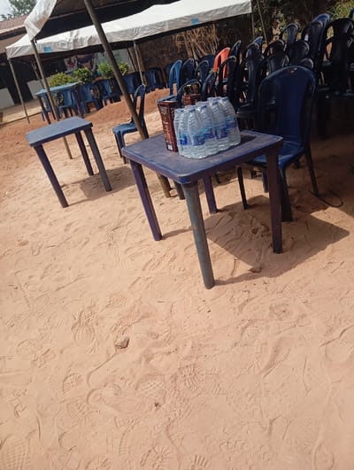 The scene depicts an outdoor gathering area located in Okpatu-Ukehe, Nigeria. The ground consists of light brown sand, visibly covered with numerous shoe and sandal footprints, indicating prior foot traffic. Overhead, a large white canopy tent is erected, supported by dark poles. Underneath and around the tent, numerous dark blue plastic chairs are arranged; some are individually placed, while others are stacked. Several small, rectangular dark blue plastic tables are present. In the foreground, one such table holds a six-pack of bottled water, branded "Okowa," alongside a brown rectangular item, possibly a carton or package. Another similar table is visible further back to the left. A dark building structure is partially visible behind the tent in the background. The area appears set up for an event or gathering. No individuals are visible in the image.