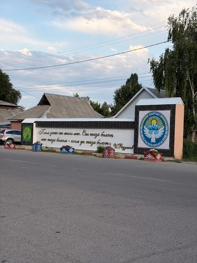 The image captures a street scene in the city of Kant, Kyrgyzstan, featuring a white wall with decorative elements and text. On the left side of the wall, a green emblem with a yellow design is visible. The wall itself displays ornate golden lettering in what appears to be Cyrillic script, along with a larger emblem on the right side. This emblem is circular, predominantly blue and yellow, depicting mountains, a sun, and a bird, with text around its perimeter. Two decorative planters with red and white patterns and filled with flowers are placed in front of the wall, one on the left and one on the right. A silver SUV is parked to the left of the wall, partially visible. The background shows a building with a grey, corrugated roof and trees, set against a sky with scattered clouds. The foreground is dominated by a paved road.