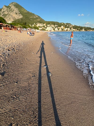 In the city of Miljevci, Montenegro, a beach scene unfolds under a clear blue sky. The foreground is dominated by a sandy and pebbled beach, with the long shadow of a person stretching across it, indicating a low sun angle, likely late afternoon or early morning.
Further down the beach, a person stands in the shallow water, facing away from the camera and towards the sea. They are wearing blue swim trunks. The water is calm, with gentle waves lapping the shore.
In the background, a row of beach umbrellas and loungers are set up, suggesting a populated area. Beyond the beach, a hillside covered in lush green vegetation rises steeply, with buildings scattered across its slopes, characteristic of a coastal town. A large, rugged mountain peak dominates the skyline in the distance. The overall impression is one of a peaceful, sunny day at the coast.
