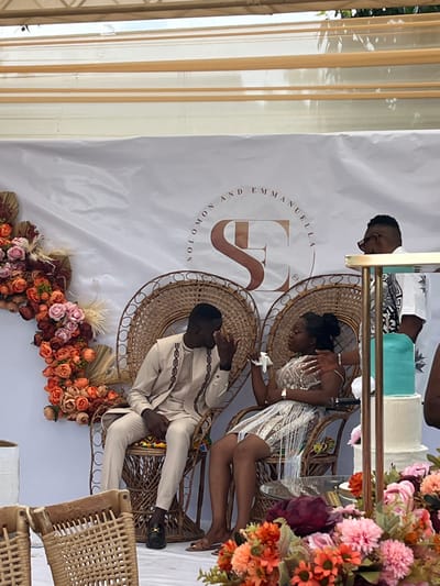 The image depicts an indoor or semi-outdoor celebratory event in Kumasi, Ghana. Two individuals, a man and a woman, are seated centrally on ornate, high-backed wicker "peacock" chairs. The man, on the left, has dark skin, wears a light beige long-sleeved tunic with dark embroidered details, matching trousers, and dark loafers. His right hand is raised to his face, near his eye, suggesting he is wiping tears or expressing strong emotion. The woman, on the right, also has dark skin and wears a shimmering, short, sleeveless dress with fringe embellishments. She holds a white tissue to her left eye/cheek, indicating a similar emotional state. A hand, likely belonging to another individual, rests gently on her left shoulder.
Behind the seated couple, a large white fabric backdrop is visible. Centered on this backdrop are the words "SOLOMON AND EMMANUELA" above a prominent, stylized "SE" monogram in a bronze tone. To the left, a large floral arrangement composed of orange, peach, pink, and maroon flowers with dried botanical elements is affixed to the backdrop. To the right of the couple, a multi-tiered cake with a teal top layer and white lower layers is partially visible, positioned behind a gold-colored metal stand. Another individual, a man with dark skin, sunglasses, and a white short-sleeved shirt with a black abstract pattern, is partially visible standing to the far right. The event space is covered by a light-colored canopy or tent structure. Additional floral arrangements are blurred in the immediate foreground.