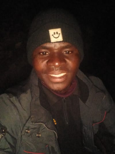 The media file is a portrait of a person, likely a young man, wearing a black beanie with a white square patch featuring a smiley face. He is also wearing a dark jacket, possibly a puffer jacket, over a dark garment with a purple collar. The background is dark, suggesting a nighttime or dimly lit setting. The person is smiling, with his teeth visible, and his eyes are open, looking towards the camera. The image is rotated, with the person's head appearing to be on the right side of the frame. Some text is visible on the jacket, which appears to be "102" in yellow lettering. The context provided indicates the location is Kwang, Nigeria. No specific weather conditions are discernible. The overall mood of the image is positive and friendly, conveyed by the smile.