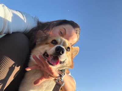 A low-angle shot shows a woman holding a corgi dog against a bright blue sky. The woman's face is visible in the lower center of the frame. She has dark hair and is wearing a light-colored shirt or sweater. Her right hand is around the dog. The corgi has tan and white fur and its tongue is hanging out. The dog wears a brown harness with a metal buckle. A portion of the woman's brown leggings or pants is visible in the upper right of the frame. The location is Gangloff, Germany.