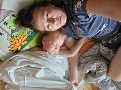 A mother and her baby are lying down on a bed. The mother is looking at the camera and smiling. The baby is asleep, with its head on the mother's chest. The baby is wrapped in a white blanket. The bed is covered with a sheet that has a cartoon frog and flower pattern. The room is decorated with a patterned blanket. The location is Буренка, Russia.