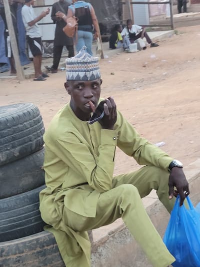 A man sits on a stack of tires in Suleja, Nigeria. He is wearing a light green traditional outfit and a patterned hat. He holds sunglasses in his right hand. He is also wearing a watch. To his right is a blue plastic bag. In the background, there are other people and buildings. The ground appears to be dirt or sand.