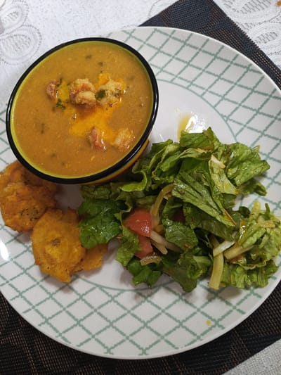 A close-up, overhead shot shows a colorful bowl of soup, two plantain tostones, and a side salad with lettuce and tomato on a white plate with a green geometric pattern. The soup is a rich, orange-brown color and appears to have shrimp in it. The tostones are golden brown and crispy. The salad is made of fresh green lettuce and chunks of red tomato, with some thinly sliced white onions visible. The plate is set on a dark placemat and a white tablecloth. The scene is indoors, likely in a restaurant or home dining area in Guayaquil, Ecuador.