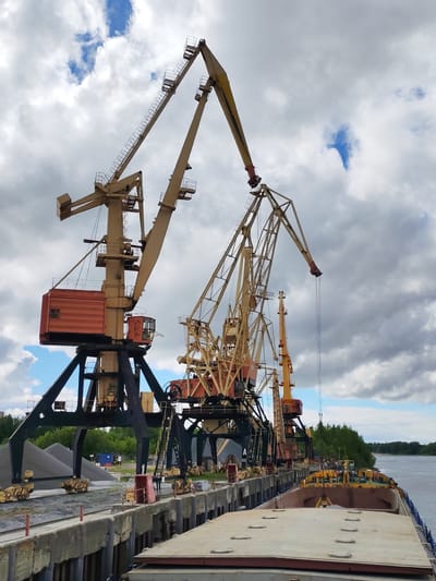 The image shows a port scene in Rybinsk, Russia, likely situated on the Volga River. Several large industrial port cranes are visible, characterized by their yellow and brown coloring and lattice-like construction. These cranes are arranged along a concrete dock, where piles of gravel are also present. A cargo barge rests alongside the dock, appearing to be loaded or unloaded with the cranes. The sky above is overcast, with numerous gray and white clouds. In the background, a strip of green vegetation indicates a riverbank.