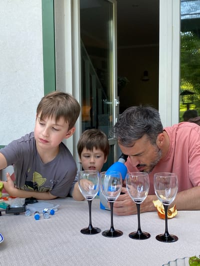 This is a photograph taken outdoors, likely on a patio or deck, with a blurred background that suggests a residential setting with trees and sunlight. The primary subjects are a man and two young boys sitting at a table covered with a striped tablecloth.

In the foreground, on the table, are several stemmed wine glasses with black bases, a blue microscope, and various small science-related items including vials with blue caps and a clear plastic case. There is also a dried piece of fruit.

The boy on the left, with brown hair, is wearing a grey t-shirt with a graphic print and is looking down towards the table. The boy in the middle, also with brown hair, is wearing a dark t-shirt and is looking forward. The man on the right, with grey-streaked dark hair and a beard, is wearing a pink t-shirt and is leaning over the table, appearing to look through the microscope. His right hand is near the microscope.

The architectural elements in the background, specifically the white door frame and window, suggest a modern home. The lighting indicates it is daytime, with natural light illuminating the scene. The overall impression is of a family engaged in an educational activity.

There is no indication in the image or context provided that this scene is located in the city of Lobnya, Russia.
