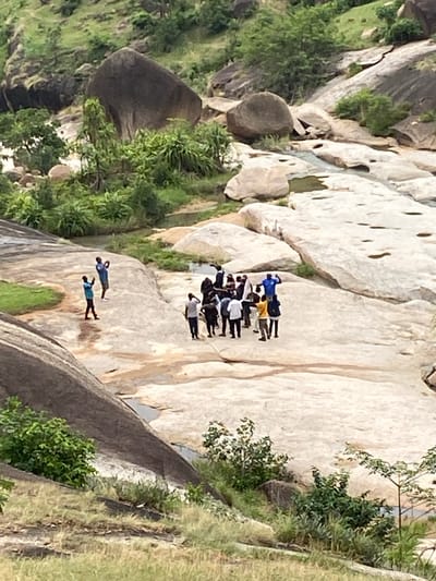 The image shows a rocky landscape in Ganawuri, Nigeria, with visible rock formations and green vegetation. A group of approximately 10-12 people are standing closely together on a large, light-colored rock surface. Below them, two individuals are lying down on the same rock formation. The scene is shot from an elevated perspective, looking down on the group and the surrounding environment.