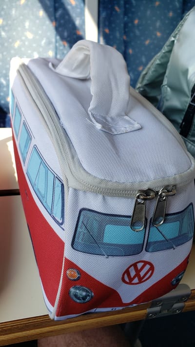The image shows a close-up view of a white cooler bag designed to resemble the front of a red and white Volkswagen Type 2 van, commonly known as a "campervan" or "bus." The bag has a white carrying handle at the top and two zipper pulls on the front. The design features stylized windows and headlights, with a Volkswagen logo prominently displayed on the front. The cooler is placed on a wooden surface, likely a table, with a blurred background that appears to be the interior of a vehicle or a room with patterned blue curtains. The lighting suggests it might be daytime, with sunlight filtering through. The scene is static and does not show any people or ongoing activities. The setting is not identifiable as Dolný Hričov, Slovakia, based solely on the visual information provided.