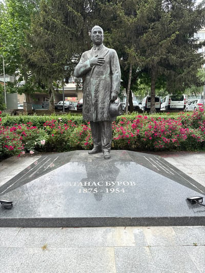 A full-body bronze statue of a male figure stands prominently on a multi-tiered, dark grey granite plinth. The statue depicts the man in a suit, tie, and long coat, with his right hand placed over his chest and his left hand holding a fedora-style hat. The plinth bears the inscription "АТАНАС БУРОВ" (Atanas Burov) and the dates "1875 - 1954" in light grey capital letters. Two small black floodlights are affixed to the plinth, positioned to illuminate the statue.
The statue and plinth are situated on a light grey tiled pavement. Immediately behind the plinth, a vibrant bed of flowering shrubs displays numerous small, bright pink blossoms, interspersed with some lighter pink or white flowers. Further back, a dense array of green foliage from various trees, including what appears to be conifers and deciduous trees, forms a natural backdrop.
In the midground and background, an urban environment is visible. Multiple white commercial vans and darker passenger vehicles are parked on what appears to be a street or parking lot. Parts of multi-story buildings with balconies are discernible beyond the trees and vehicles. A streetlamp pole is present on the left, and a green refuse bin is visible near the parked cars. The scene takes place in a public outdoor area within the city of Gorna Oryahovitsa, Bulgaria.