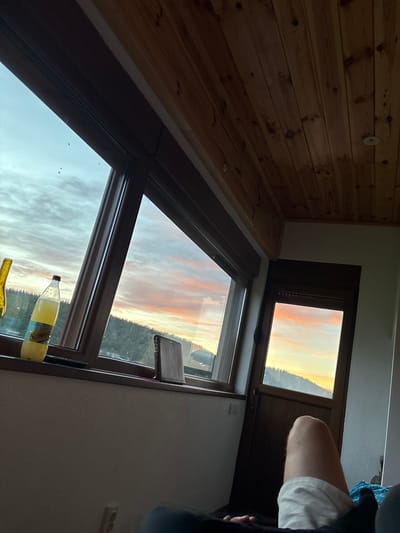 The image captures a serene view of a sunset over a forested landscape, observed from inside a room with a wooden paneled ceiling. Large windows offer a panoramic vista, with the sky painted in hues of orange, pink, and blue. On the windowsill of the larger window, a bottle of "Schweppes" sparkling lemonade is placed next to a yellow bottle. A dark, rectangular object, possibly a book or a wallet, rests on the sill as well. To the right, a smaller window or doorframe reveals a similar sunset view. The lower portion of the image shows a person's leg, clad in white shorts, resting on what appears to be a bed. The overall ambiance suggests a moment of relaxation and appreciation of nature's beauty. This scene is not located in Virak, Montenegro.