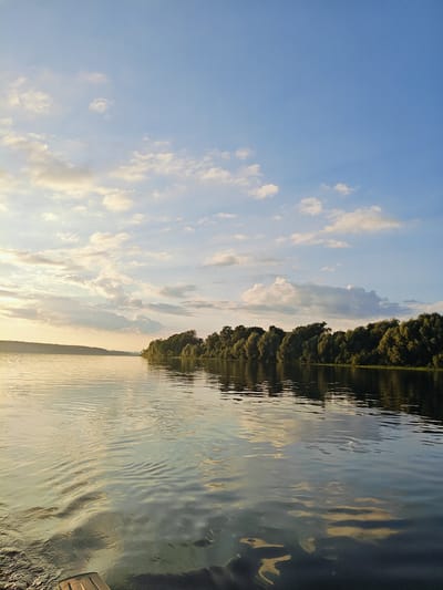 This media file depicts a serene natural landscape at dusk, likely in Chernushka, Russia. The scene is dominated by a wide, calm body of water reflecting the soft, warm light of the setting sun. A dense line of lush green trees forms a natural border along the opposite bank, creating a striking contrast with the water's surface. Above, the sky transitions from a soft blue to warmer hues, scattered with fluffy white clouds that catch the fading sunlight.
The time of day is clearly evening, indicated by the golden light and long shadows, suggesting a peaceful close to the day. The weather appears pleasant, with clear skies and calm water, ideal for enjoying the outdoors. The overall emotion evoked is one of tranquility and natural beauty, with a sense of stillness and quietude. There are no visible people or text in the media file. The image captures a moment of natural splendor, emphasizing the harmonious relationship between the water, the trees, and the sky.