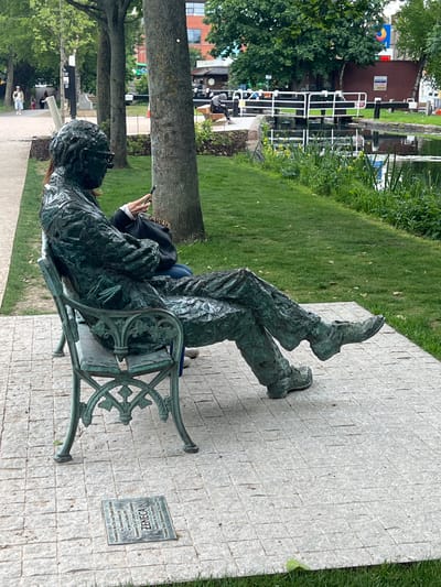 A statue of a person lying on a bench is situated outdoors next to a walkway and grassy area. The statue is near a plaque that reads "Zeneca" and gives further details in a small font. A person in a black jacket is sitting on the same bench. A body of water with locks is visible in the background. The scene appears to be located in Dublin, Ireland.