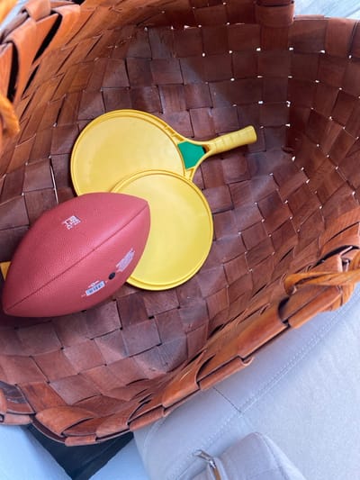 Inside a woven, brown basket are two yellow beach paddle ball rackets and a red-brown American football. The rackets are overlapping each other. The football lies near the paddles, slightly covering one. The basket's weave pattern is visible, adding texture. A portion of a light-colored, upholstered couch is visible in the upper right corner.