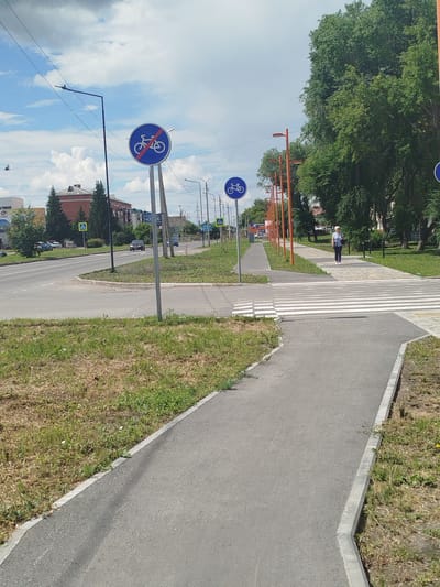 A street scene in Zavodoukovsk, Russia, captured on a sunny day with a partly cloudy sky. The image features a paved road with a pedestrian crossing, a bicycle path, and a sidewalk. Several signs are visible, including a "No cycling" sign and a pedestrian crossing sign. A woman is walking on the sidewalk, and cars are present on the road. Lush green trees line the right side of the image, with buildings visible in the background. The overall impression is of a well-maintained urban environment with infrastructure for both vehicles and pedestrians.