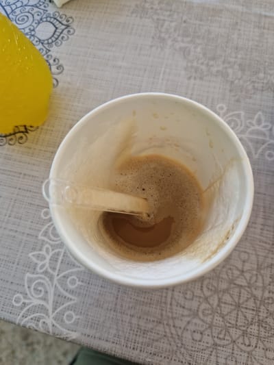 A close-up, overhead shot shows a white paper cup containing coffee with foam. A clear plastic stirrer is placed across the rim of the cup, partially submerged in the coffee. The cup rests on a table covered with a light gray tablecloth that features a subtle white floral or paisley pattern. To the left of the cup, a bright yellow object, possibly a piece of fruit or a drink, is partially visible. The overall scene suggests a casual setting, likely indoors at a cafe or restaurant, not specifically located in German, Bulgaria.