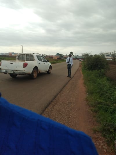 A white pickup truck drives on a paved road under a cloudy sky. A man wearing a white dress shirt and dark pants walks on the side of the road. The road is lined with grass and trees. In the background, there are buildings and houses. The scene appears to be in Du, Nigeria, a city known for its rural setting and lush vegetation.