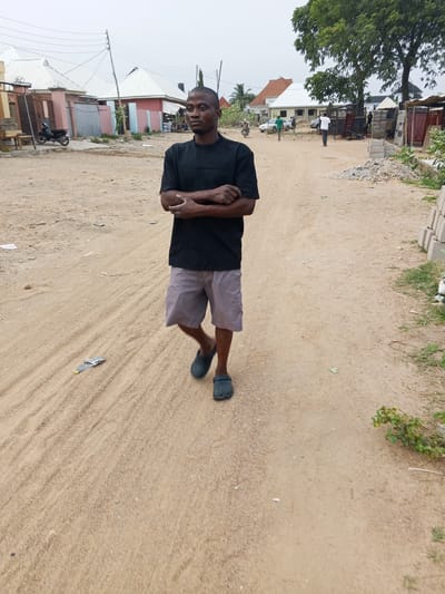 A medium shot captures a man standing in a sandy road in Otokiti, Nigeria. He has short dark hair, a black T-shirt, grey shorts, and dark blue footwear. His arms are crossed. The road stretches into the distance, flanked by buildings, trees, and vegetation. The sky appears overcast. In the background are buildings with white or red roofs, power lines, and other figures moving further down the road. To the right of the frame are piles of grey construction blocks and scattered vegetation.