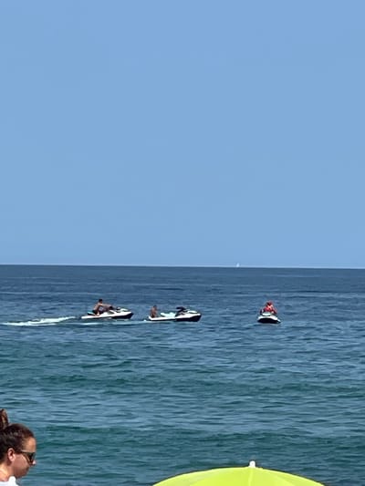 The image captures a coastal scene with the vast expanse of the Mediterranean Sea under a clear blue sky, likely near Badalona, Spain. Three jet skis are the prominent subjects on the water. The first jet ski, to the left, is ridden by a person with dark hair, leaving a white wake. To its right, a second jet ski is occupied by two individuals, one of whom is leaning forward. Further to the right, a third jet ski carries a person wearing an orange life vest. In the distance, a faint white sail can be seen on the horizon. In the foreground, a portion of a person's head and shoulder, along with sunglasses and an umbrella, are visible, suggesting a beach setting. The water is a deep blue, transitioning to a lighter turquoise closer to the shore.