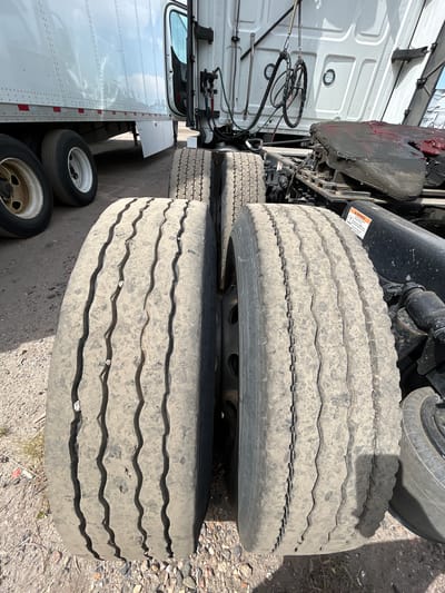 This is a close-up, eye-level, outdoor shot of the rear tires of a semi-trailer truck parked on a gravel surface. The truck has a white cab and a white trailer. The tires are heavily worn, with deep tread grooves. The right front tire of the rear axle is in focus and is slightly obscured by the left front tire of the rear axle. The left front tire is also in focus and partially obscured by the right front tire. The background shows more of the truck's undercarriage, including the fifth wheel assembly, and parts of the trailer. The sky is visible in the upper left corner. The overall impression is of a commercial vehicle in a lot or industrial area. The image does not contain any specific landmarks or details that would allow for precise location identification within Aurora, United States of America.